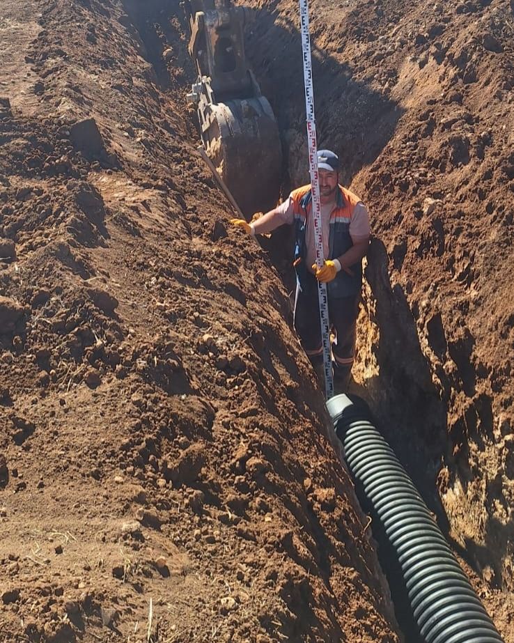 Turuncu yelekli bir işçi, kazılmış toprak kanal içinde ölçüm çubuğu tutarken siyah körüklü boru uzanıyor.