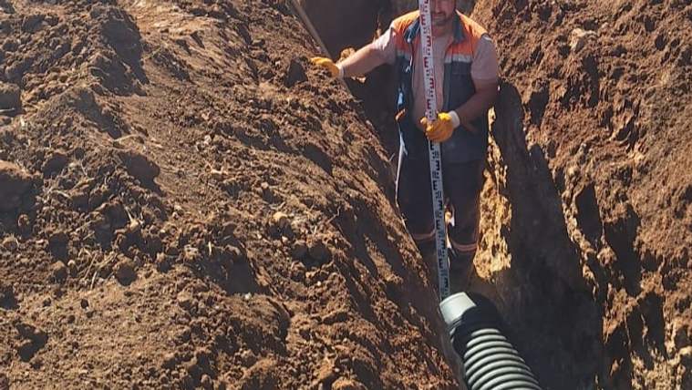 Turuncu yelekli bir işçi, kazılmış toprak kanal içinde ölçüm çubuğu tutarken siyah körüklü boru uzanıyor.
