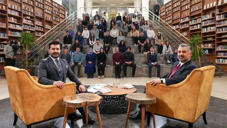 Kütüphane salonunda iki erkek koltuklarda oturuyor; arkalarında merdiven basamaklarına dizilmiş bir grup bulunuyor.