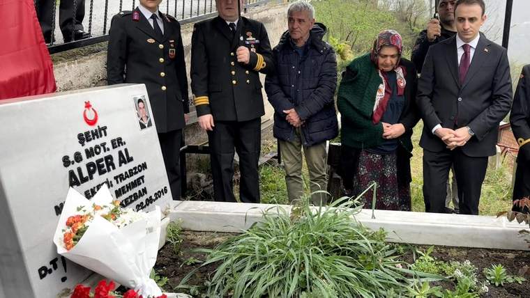 Yağmur altında bir mezar taşı, çiçekler ve kırmızı karanfiller önünde duran beş kişi görülüyor.