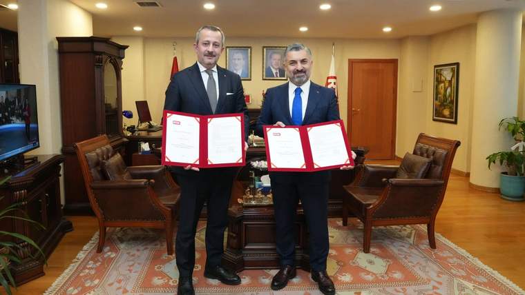 Bir ofiste iki erkek, imzalı belgelerin bulunduğu açık dosyaları kameraya doğru tutuyor.