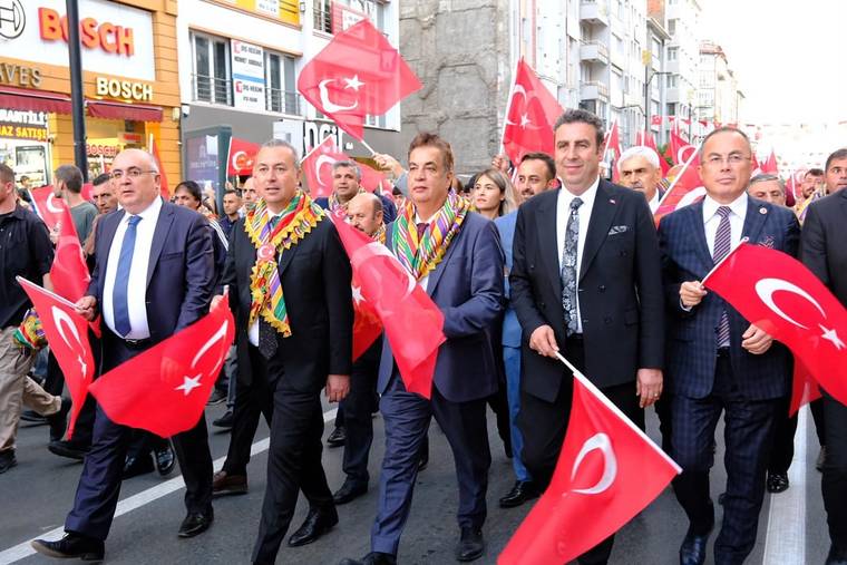 Türk bayrakları taşıyan kalabalık, kent merkezindeki caddede yürürken önde takım elbiseli kişiler görülüyor.