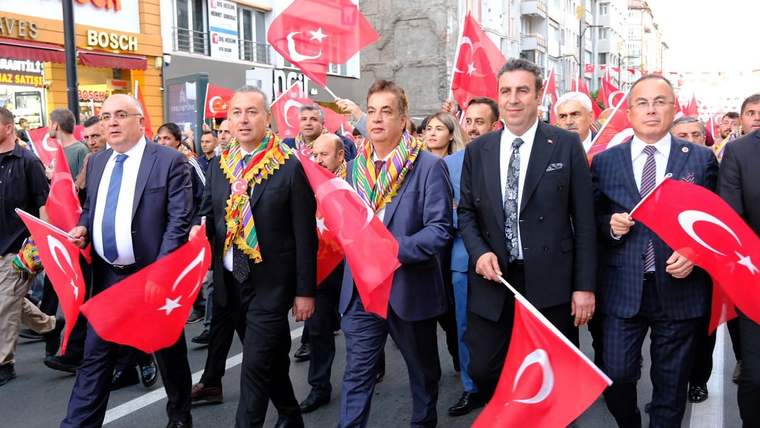Türk bayrakları taşıyan kalabalık, kent merkezindeki caddede yürürken önde takım elbiseli kişiler görülüyor.