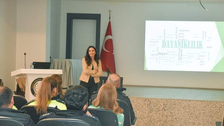 Bir salonda sunum yapan kadın, Türk bayrağı ve ekranda “DAYANIKLILIK” yazılı bir slayt görülüyor.