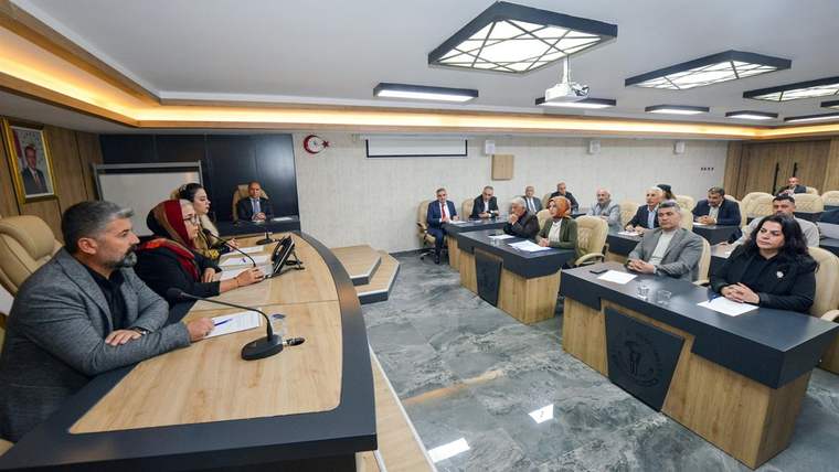 Toplantı salonunda, masada oturan beş kişi ve arka planda diğer katılımcılar yer alıyor.