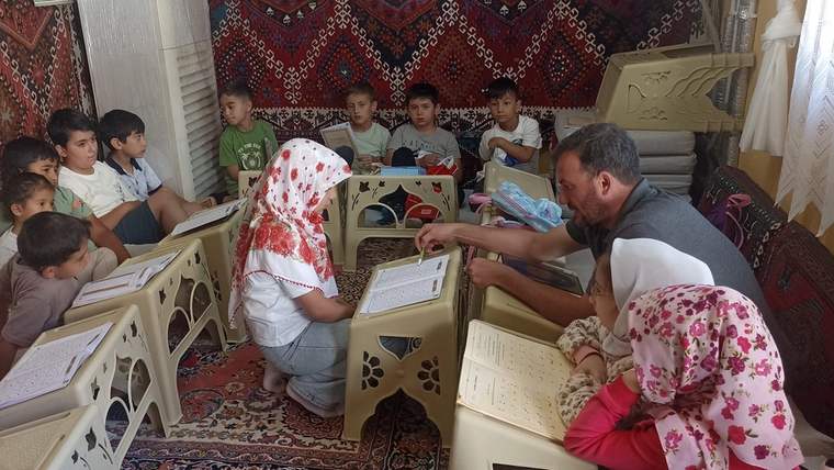 Çocuklar, bir erkek ve başörtülü bir kadın halı döşeli odada alçak masalar çevresinde kitap okuyor.