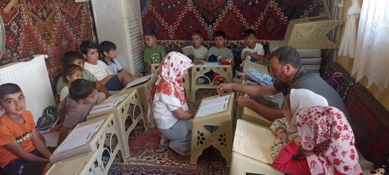 Çocuklar, bir erkek ve başörtülü bir kadın halı döşeli odada alçak masalar çevresinde kitap okuyor.