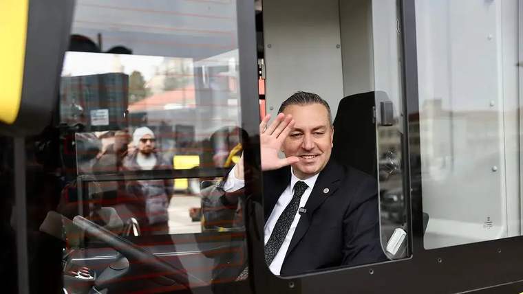 Siyah takım elbiseli bir adam, sarı-siyah otobüsün sürücü kabininden el sallıyor.