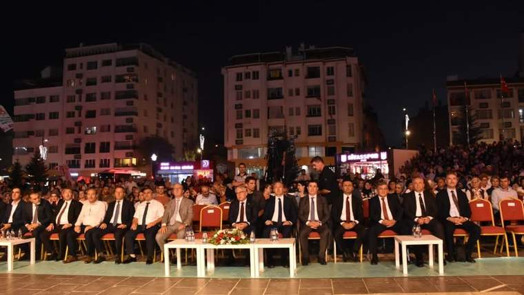 Gece düzenlenen açık hava etkinliğinde takım elbiseli kişiler sandalyelerde oturuyor, arkalarında kalabalık bulunuyor.