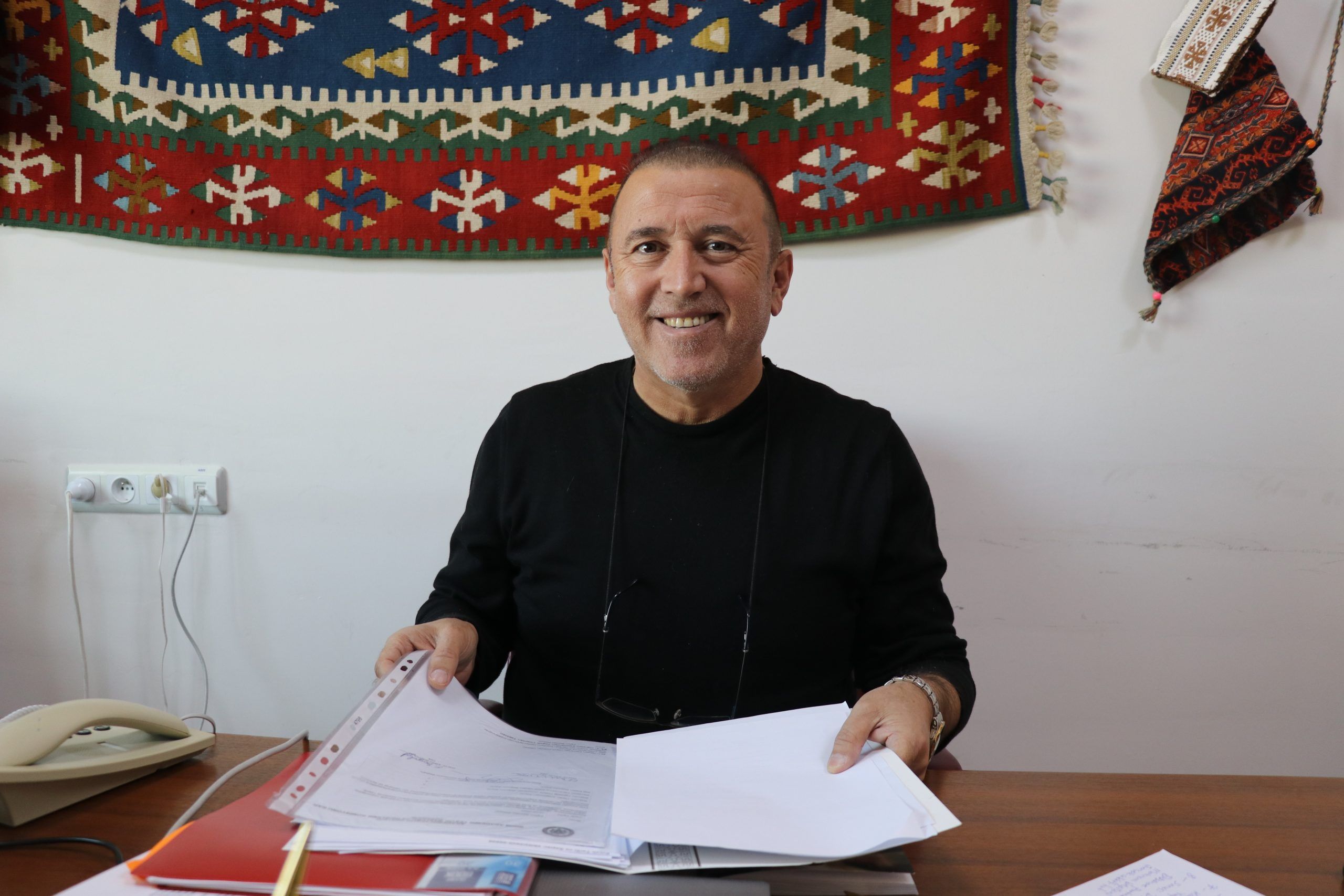 Masada oturan bir kişi, belgeleri tutarak gülümsüyor. Arka planda geleneksel bir halı asılı.