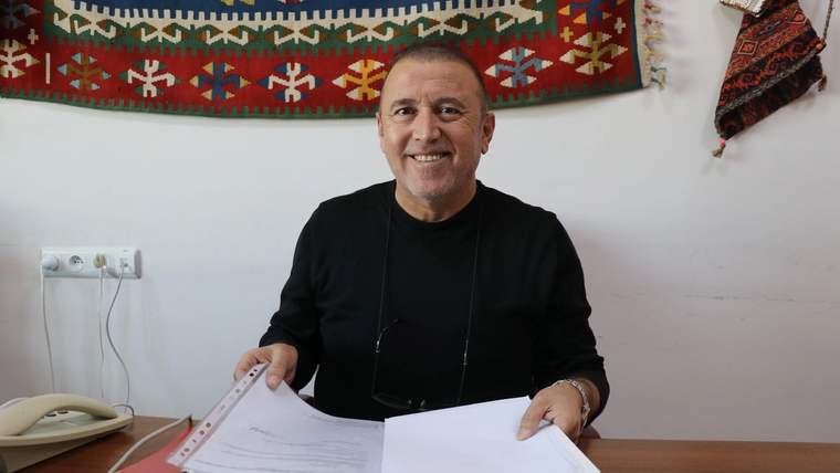 Masada oturan bir kişi, belgeleri tutarak gülümsüyor. Arka planda geleneksel bir halı asılı.