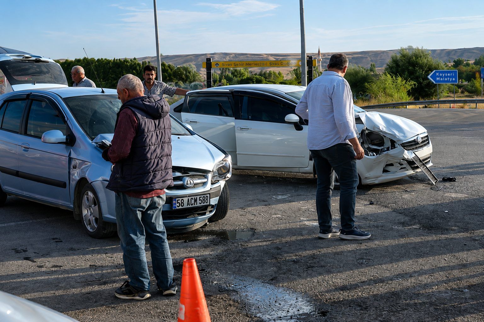 Üç otomobilin bulunduğu kazada, ön kısmı hasarlı beyaz aracın yanında insanlar ve turuncu trafik konisi görülüyor.