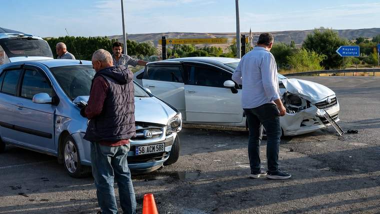 Üç otomobilin bulunduğu kazada, ön kısmı hasarlı beyaz aracın yanında insanlar ve turuncu trafik konisi görülüyor.