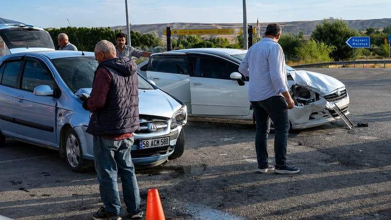 Üç otomobilin bulunduğu kazada, ön kısmı hasarlı beyaz aracın yanında insanlar ve turuncu trafik konisi görülüyor.