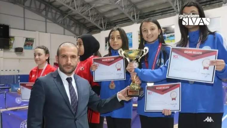 Masa tenisi salonunda beş çocuk, ellerindeki kupaları ve sertifikaları gösterirken poz veriyor.