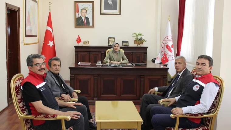 Bir masa etrafında oturan dört kişi ve arka planda bir kadın, Türk bayrağı ve tabela görünmekte.