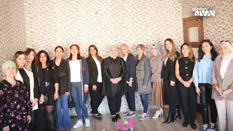 Bir salonda farklı yaşlardan kadınlar, duvar önünde yan yana ve elleri önde bağlı duruyor.