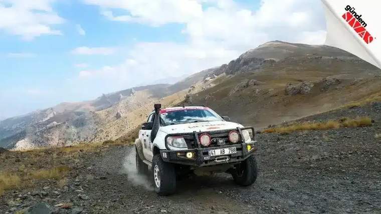 Sert arazide ilerleyen beyaz bir off-road aracı, dağlık bir manzara önünde yer alıyor.