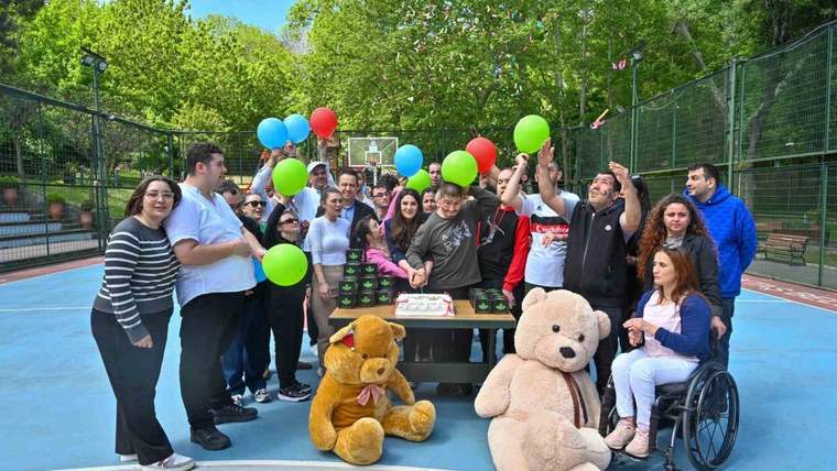 Spor sahasında balonlar, pasta ve pelüş oyuncaklarla poz veren bir grup insan; önde tekerlekli sandalye var.