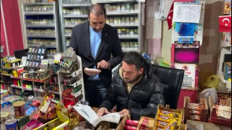 Bir dükkanda, bir kişi kitap okurken diğeri cep telefonuyla ona bakıyor. Etrafında çeşitli ürünler var.