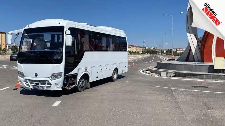 Beyaz bir minibüs, yolun ortasında duruyor. Arka planda bir heykel görünmekte.
