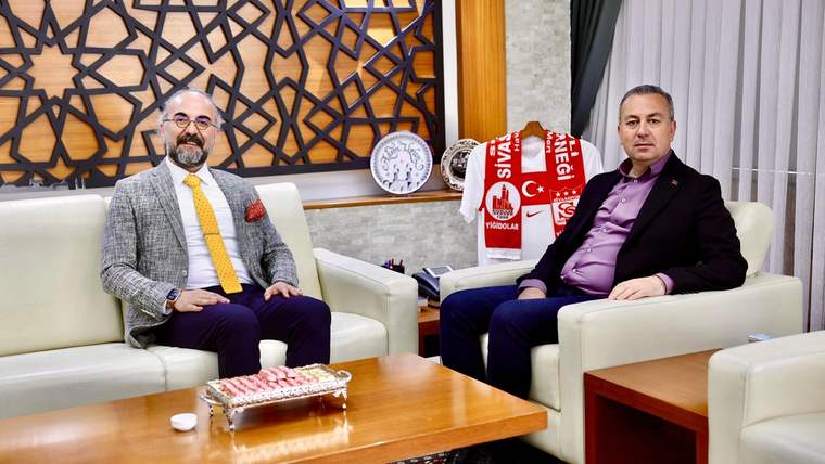 İki kişi, bir ofis ortamında oturuyor; biri gri ceket, diğeri mor gömlek giymiş.