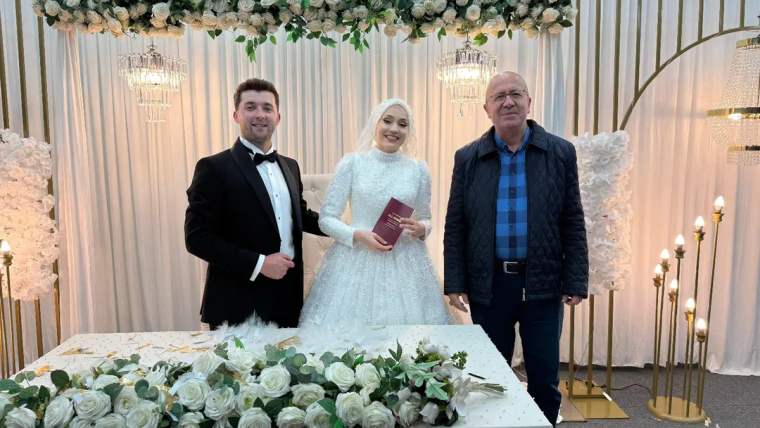 Düğün töreninde gelin ve damat, masanın önünde poz veriyor. Damat smokin giymiş, gelin beyaz elbise giymiş.