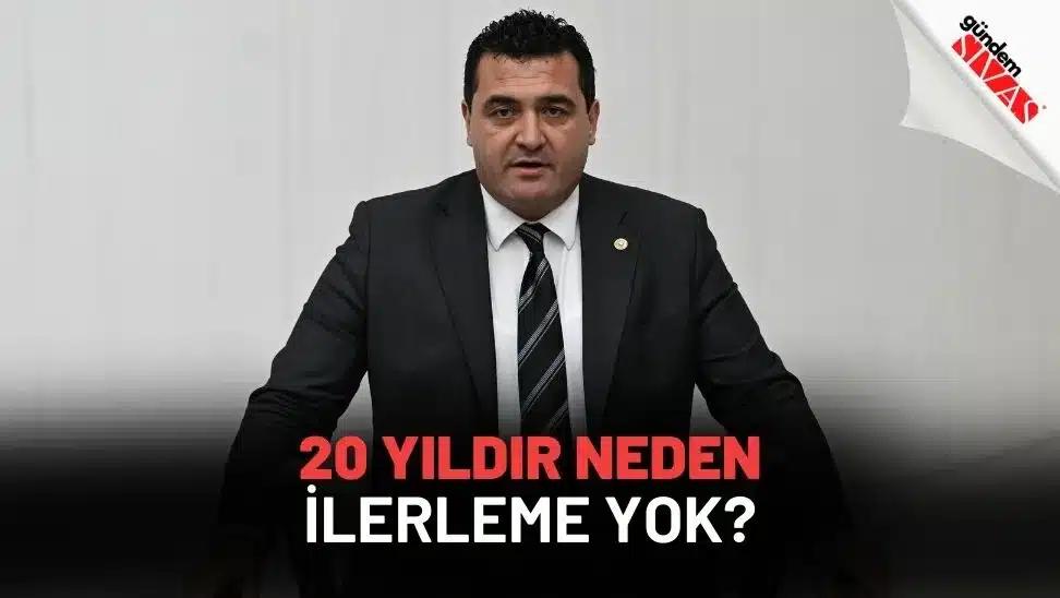 Siyah takım elbiseli bir kişi, beyaz arka plan önünde oturuyor; altta soru biçiminde yazı yer alıyor.