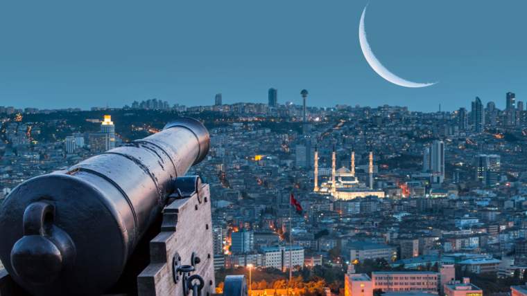 Ön planda tarihi bir top, arka planda şehir silueti, aydınlatılmış cami ve gökyüzünde hilal görülüyor.
