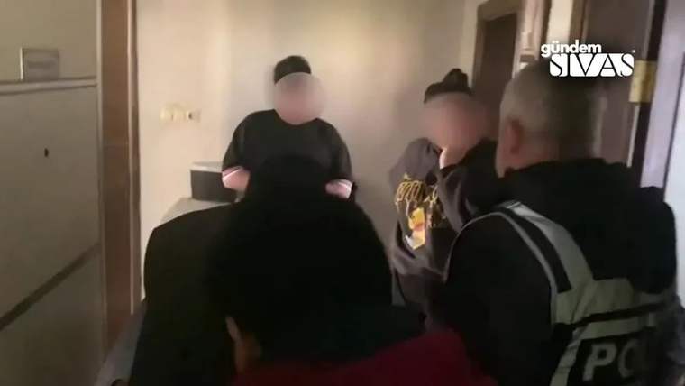 Bir koridorda yüzleri bulanıklaştırılmış birkaç kişi duruyor; sağdaki kişinin üzerinde yansıtıcı şeritli yelek var.
