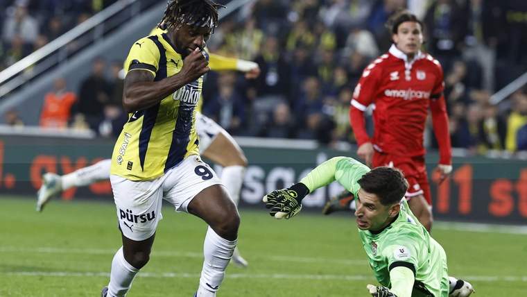 Sarı-lacivert formalı futbolcu top sürerken, yeşil formalı kaleci yerde uzanıyor; kırmızılı oyuncu arkada duruyor.