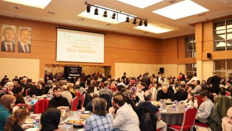 Büyük bir salonda yemek masalarında oturan kalabalık topluluk, sahne ve üzerinde “Hoş Geldiniz” yazılı perde görülüyor.