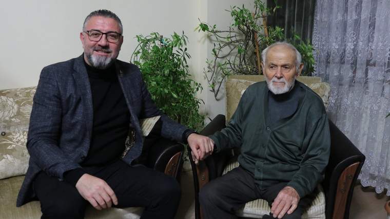 Bir kişi, yaşlı bir adamın elini tutarak oturuyor; arka planda yeşil bitkiler var.