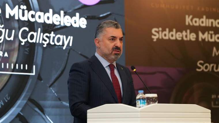 Kürsüde konuşan bir kişi, önünde su şişesi ve mikrofon var.