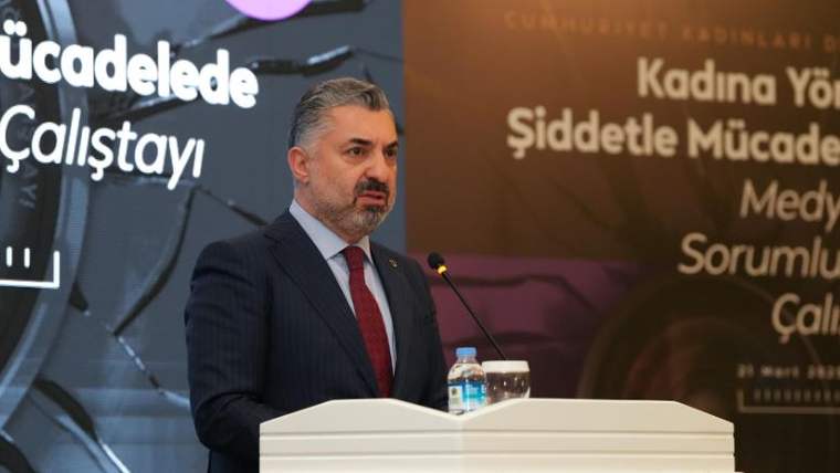 Kürsüde konuşan bir kişi, önünde su şişesi ve mikrofon var.