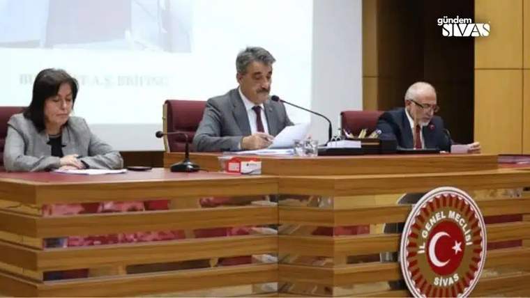 Bir toplantı masasında oturan üç kişi, belgeleri inceliyor. Arkada Türk bayrağı ve sunum ekranı var.