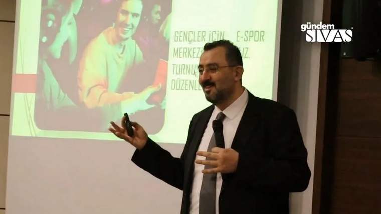 Kürsü önünde konuşan takım elbiseli bir kişi, arkasındaki ekranda e-spor sunumuyla görülüyor.