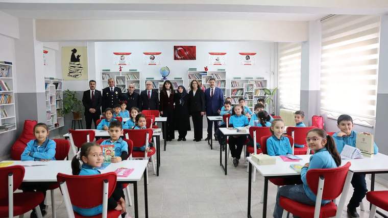 Kütüphanede, masalarda oturan çocuklar ve arka planda yetişkinler yer alıyor.