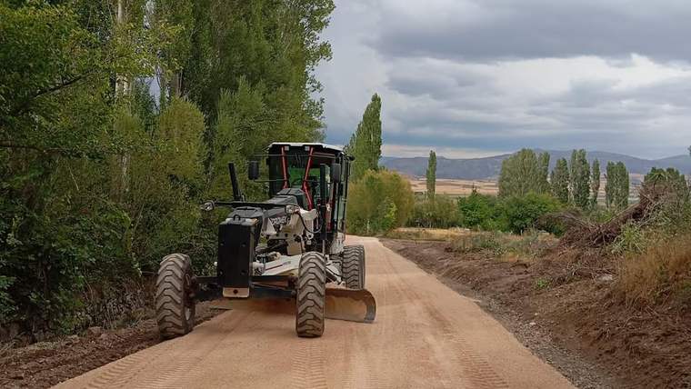 Ağaçlarla çevrili toprak yolda bir greyder, yol yüzeyini düzleştirirken görülüyor.
