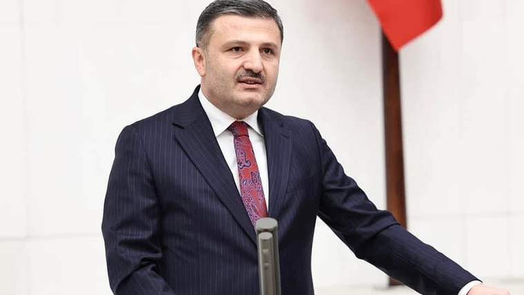 Kürsüde konuşan bıyıklı bir erkek, koyu renk takım elbise ve kırmızı kravat takıyor; arkasında Türk bayrağı bulunuyor.