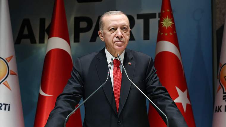 Kürsüde konuşan bir kişi, iki Türk bayrağı ve arkasında AK Parti yazılı bir fon önünde duruyor.