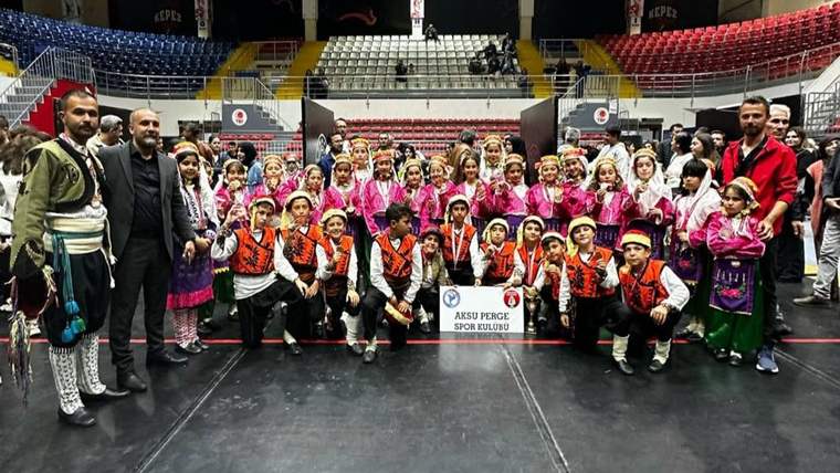 Geleneksel kıyafetler giyen çocuk ve yetişkinlerden oluşan grup, salonda Aksu Perge Spor Kulübü pankartı taşıyor.