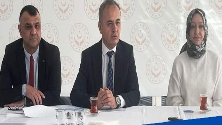 Masa başında oturan iki erkek ve başörtülü bir kadın, logolu bir duvar önünde toplantı yapıyor.
