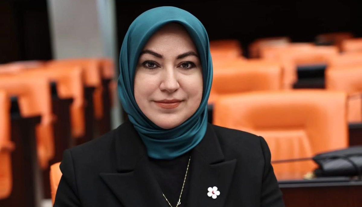 Başında başörtüsü olan bir kadın, arka planda turuncu koltuklar bulunan bir mekanda gülümseyerek poz veriyor.