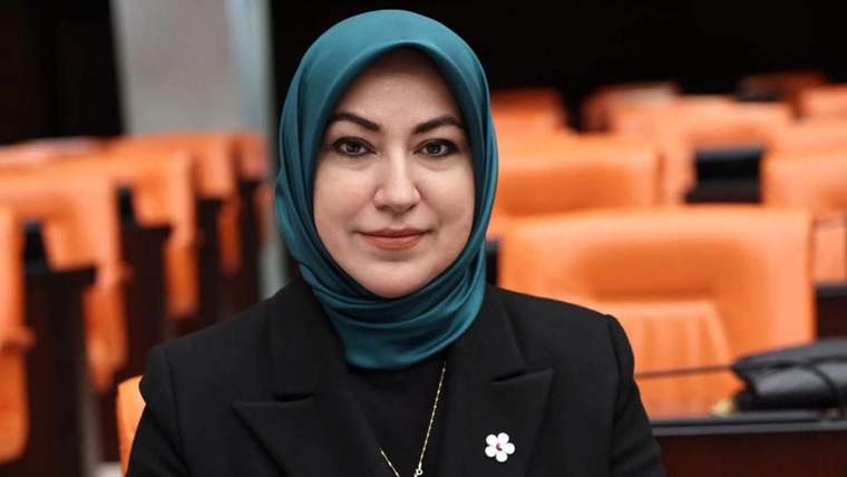 Başında başörtüsü olan bir kadın, arka planda turuncu koltuklar bulunan bir mekanda gülümseyerek poz veriyor.