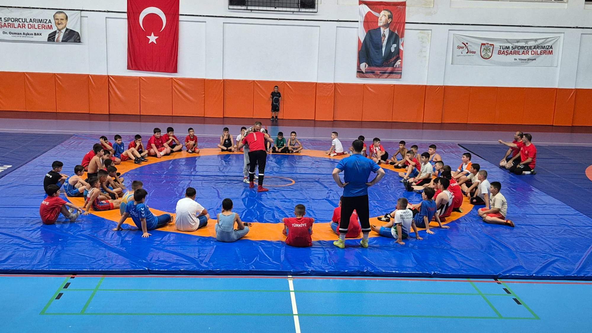 Güreş salonunda çocuklar mavi minder üzerinde daire oluştururken ortada duran iki kişi görülüyor.