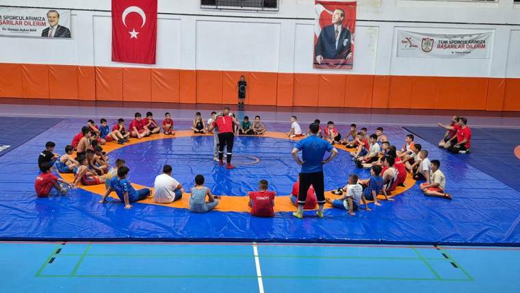 Güreş salonunda çocuklar mavi minder üzerinde daire oluştururken ortada duran iki kişi görülüyor.