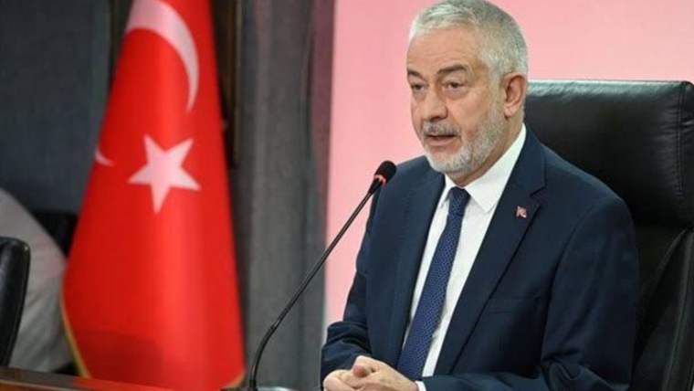 Kravatlı bir kişi mikrofonda konuşuyor; yanında Türk bayrağı ve arkasında pembe duvar bulunuyor.