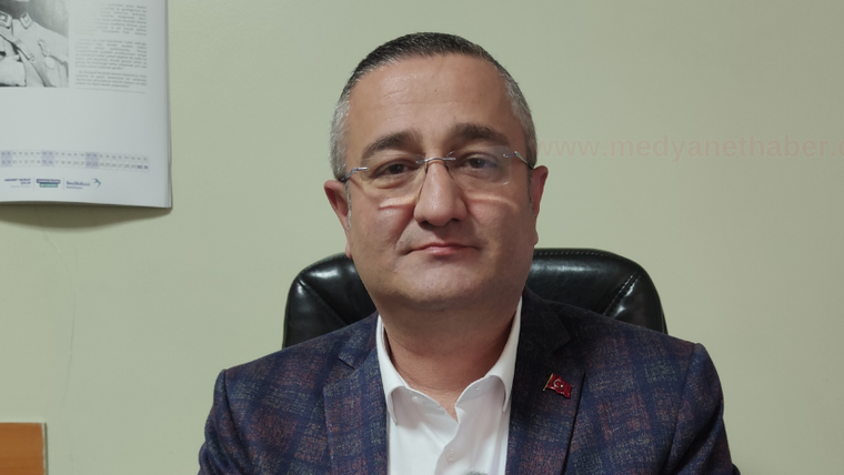 Gözlüklü bir erkek, Türk bayrağı rozeti bulunan ceketiyle ofiste koltukta oturuyor.