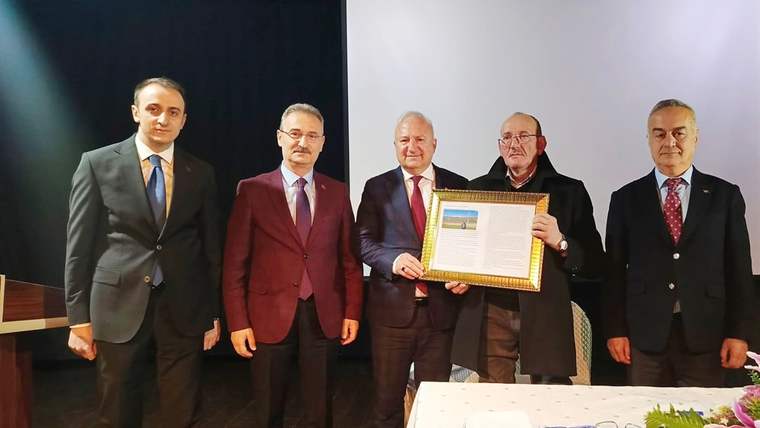 Beş erkek sahnede yan yana duruyor; ortadaki iki kişi çerçeveli bir belgeyi birlikte tutuyor.